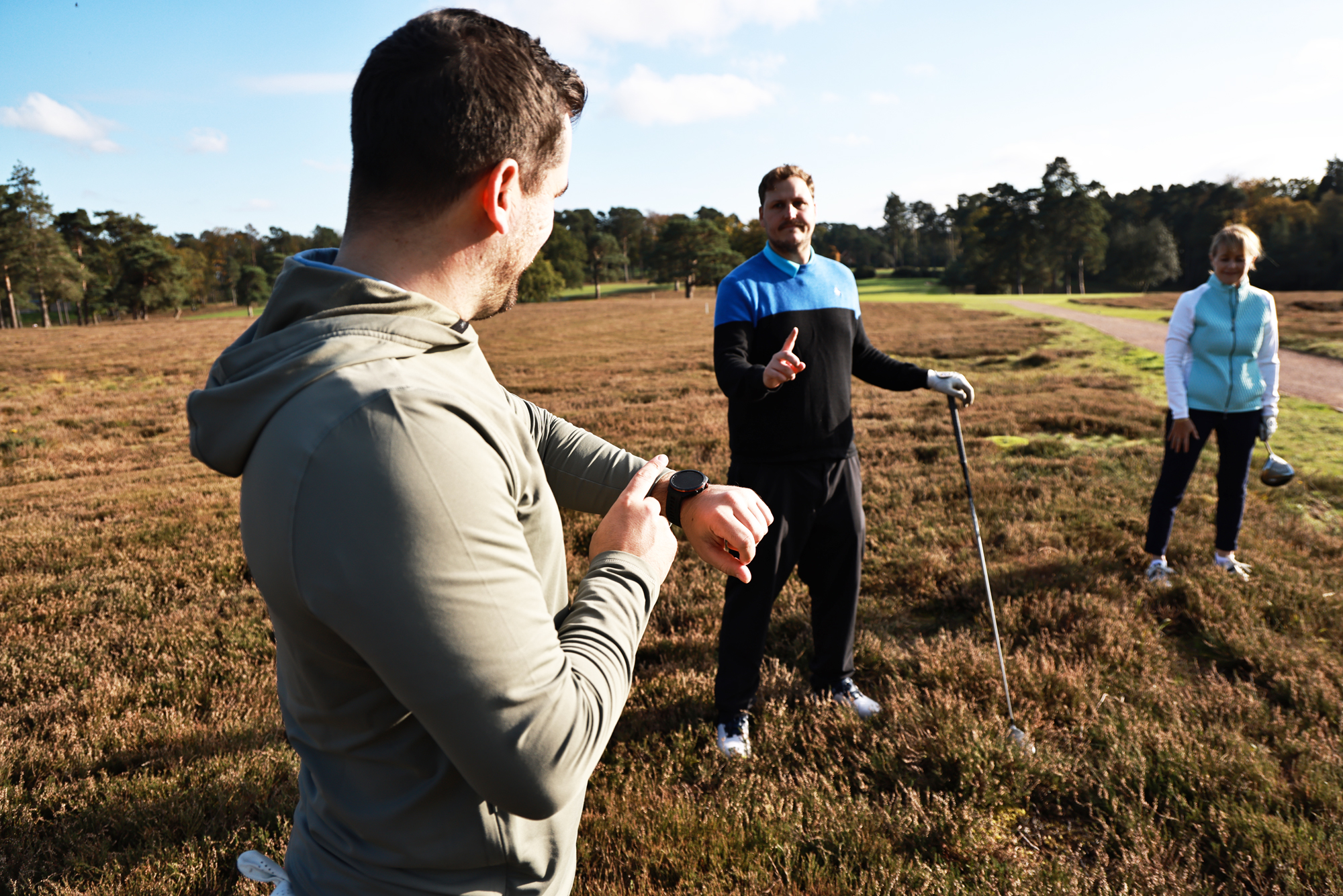 Sam De'Ath pointing to his watch as Nick Bonfield looks for his golf ball, with Alison Root also helping in the search at West Hill Golf Club