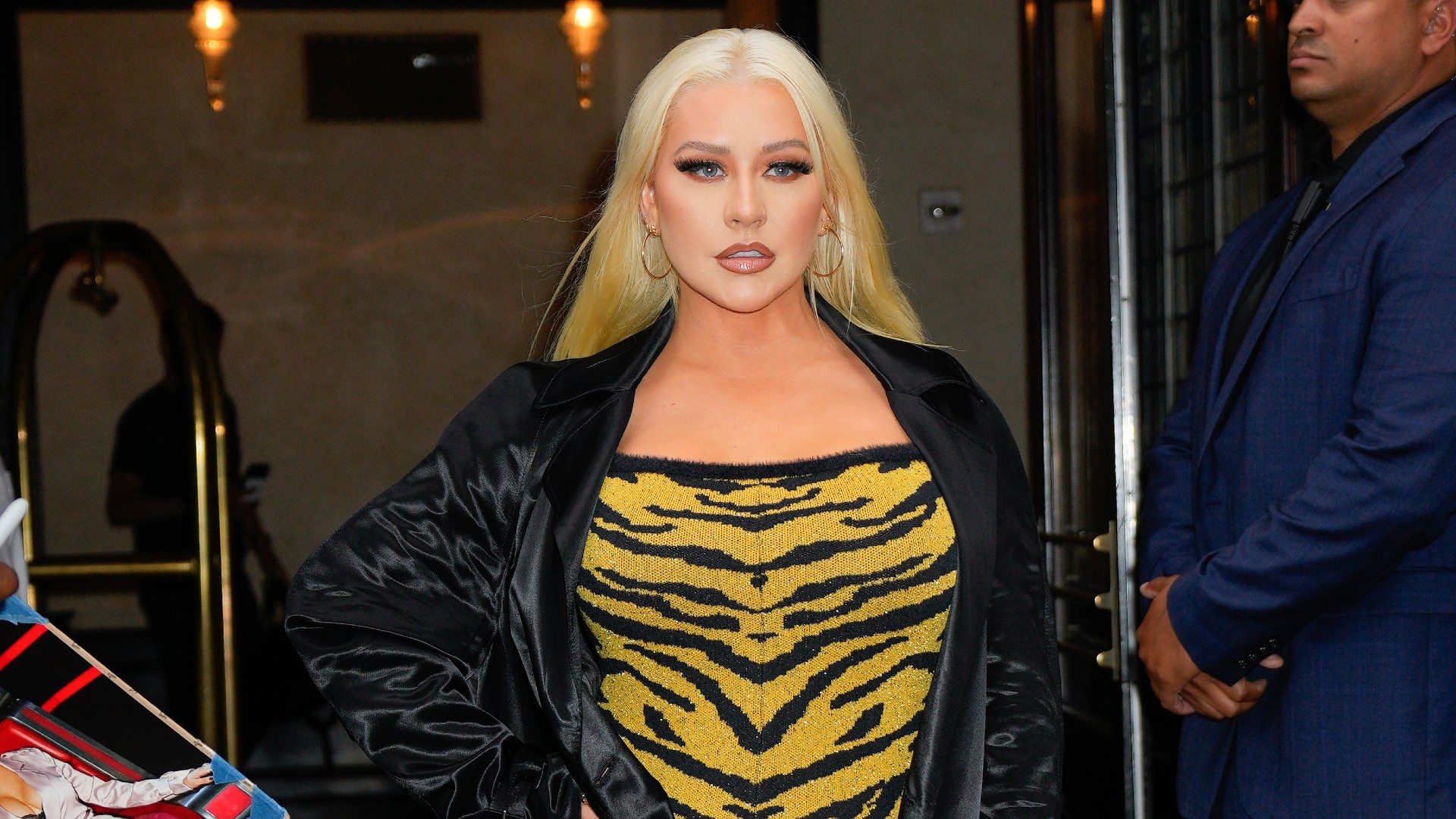 a white woman with platinum blonde hair being photographed in front of a building wearing a yellow animal print dress and a black silk jacket