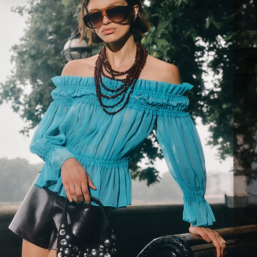 A woman modelling a blue off-the-shoulder ruffle shirt with a black PVC mini skirt and chunky beaded necklace from River Island