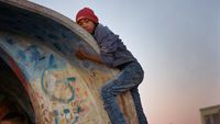 A boy in a red beanie, gray hoodie, and jeans climbs over the curved roof of a brightly graffitied structure at a bus stop during a dusky sunset