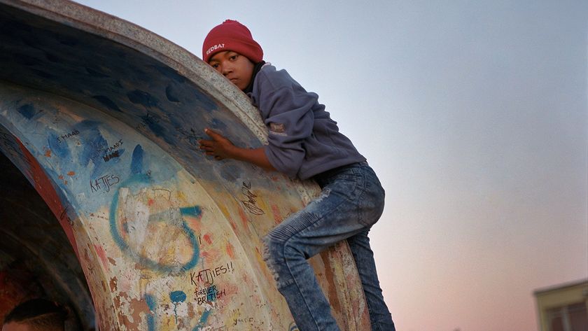 A boy in a red beanie, gray hoodie, and jeans climbs over the curved roof of a brightly graffitied structure at a bus stop during a dusky sunset