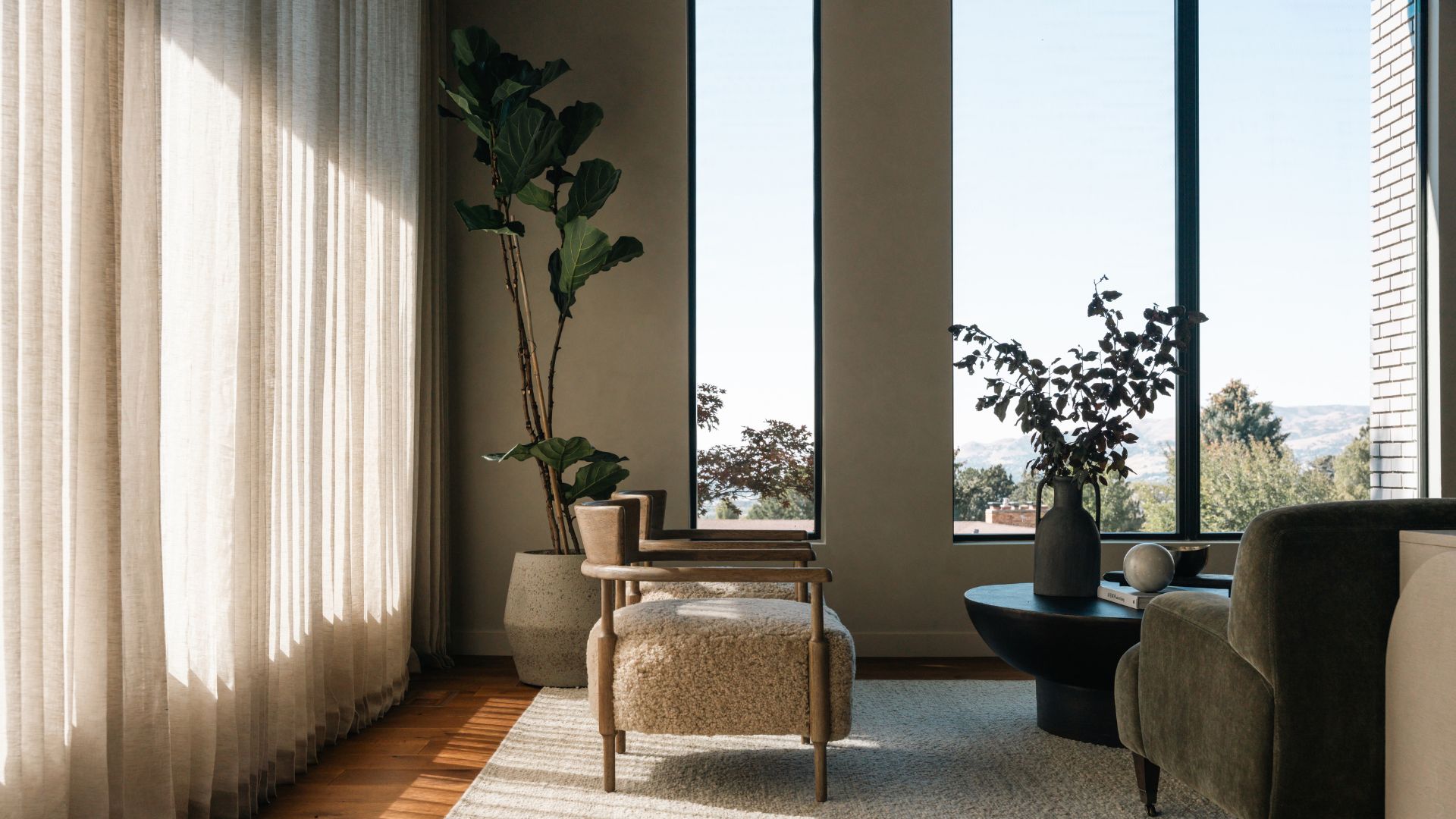 A large living room with cozy armchairs, a vase of stems, large windows, and a tall fiddle leaf fig plant