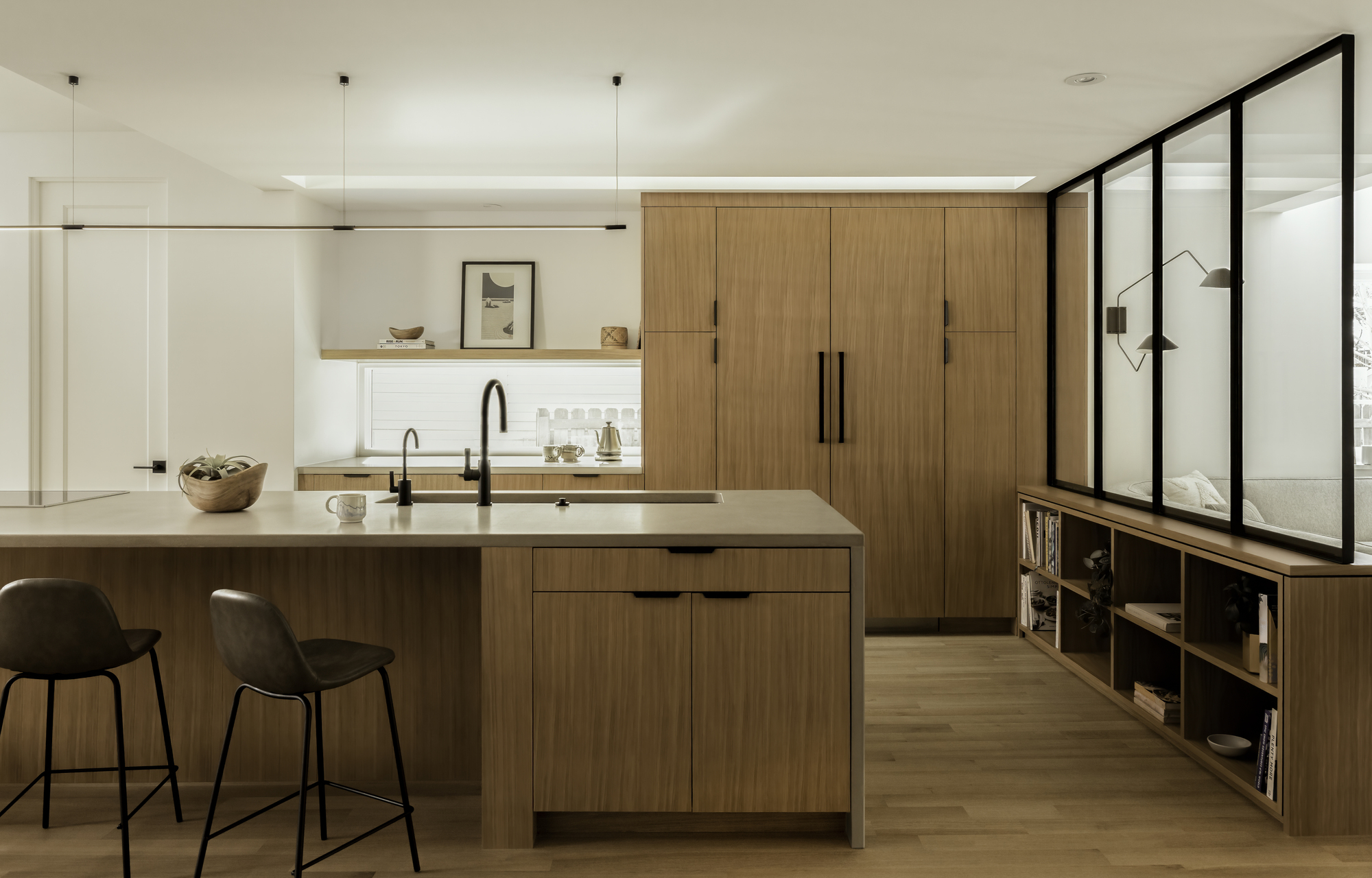 a wood kitchen with glass walls, island bench with two bar stools and a sink, bookcase, white walls, and floating timber shelves