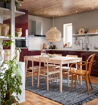 Burgundy kitchen cabinets with light wood dining table and chairs, and large natural weave pendant over table.