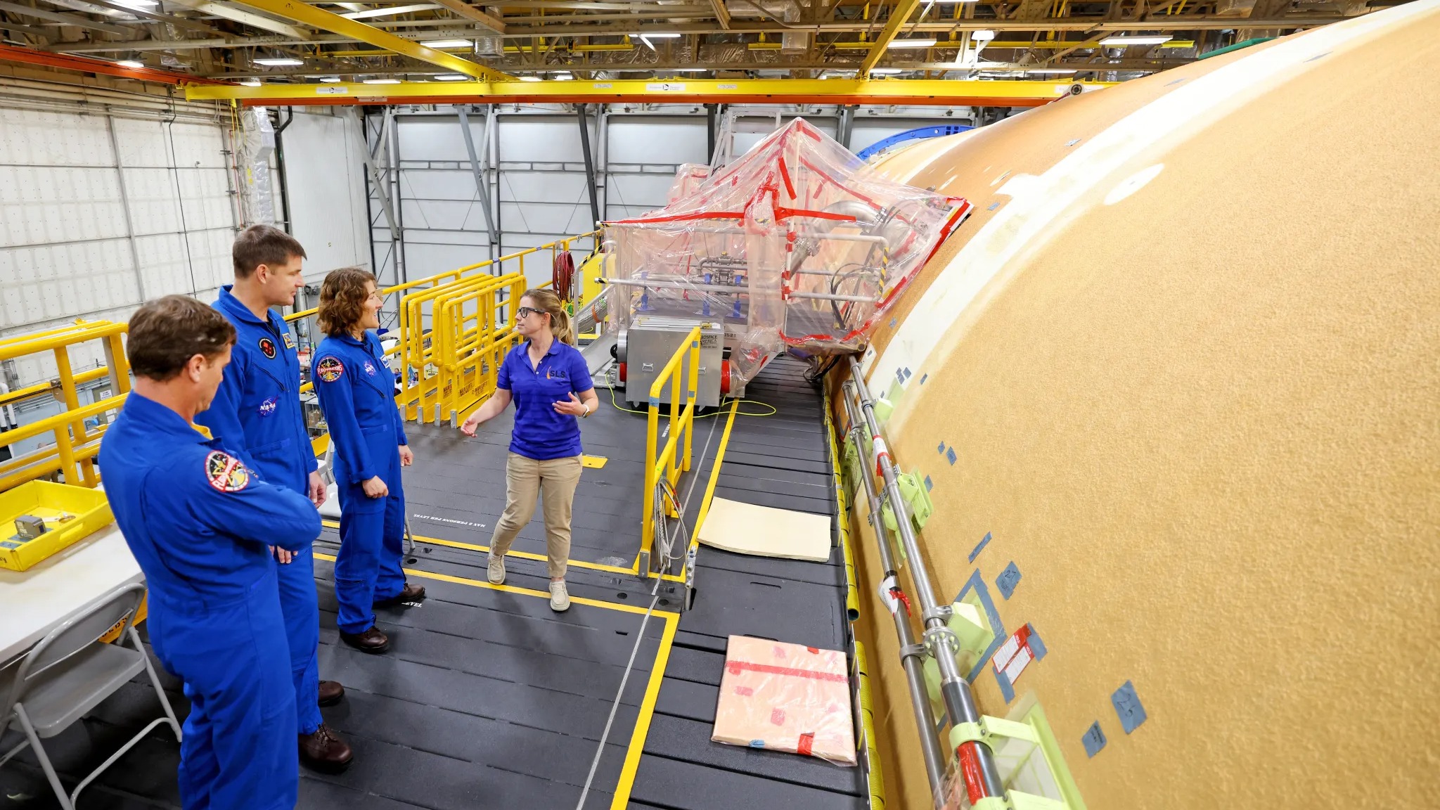 three astronauts in flight suits standing alongside a horizontal core stage of a big rocket, on a scaffolding