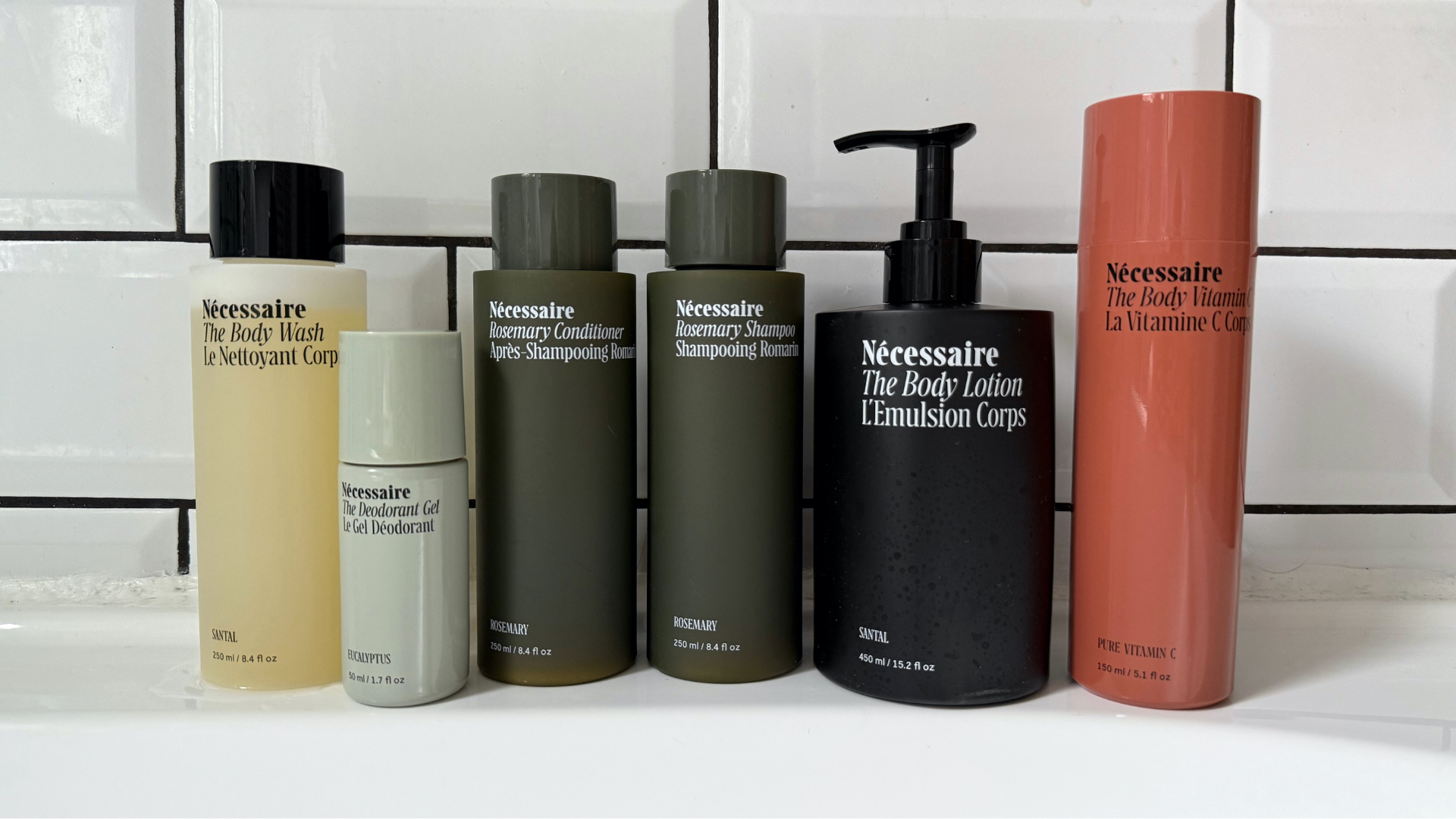 A selection of six Necessaire products stood on a white bathtub edge in front of a white tile and black grouting background