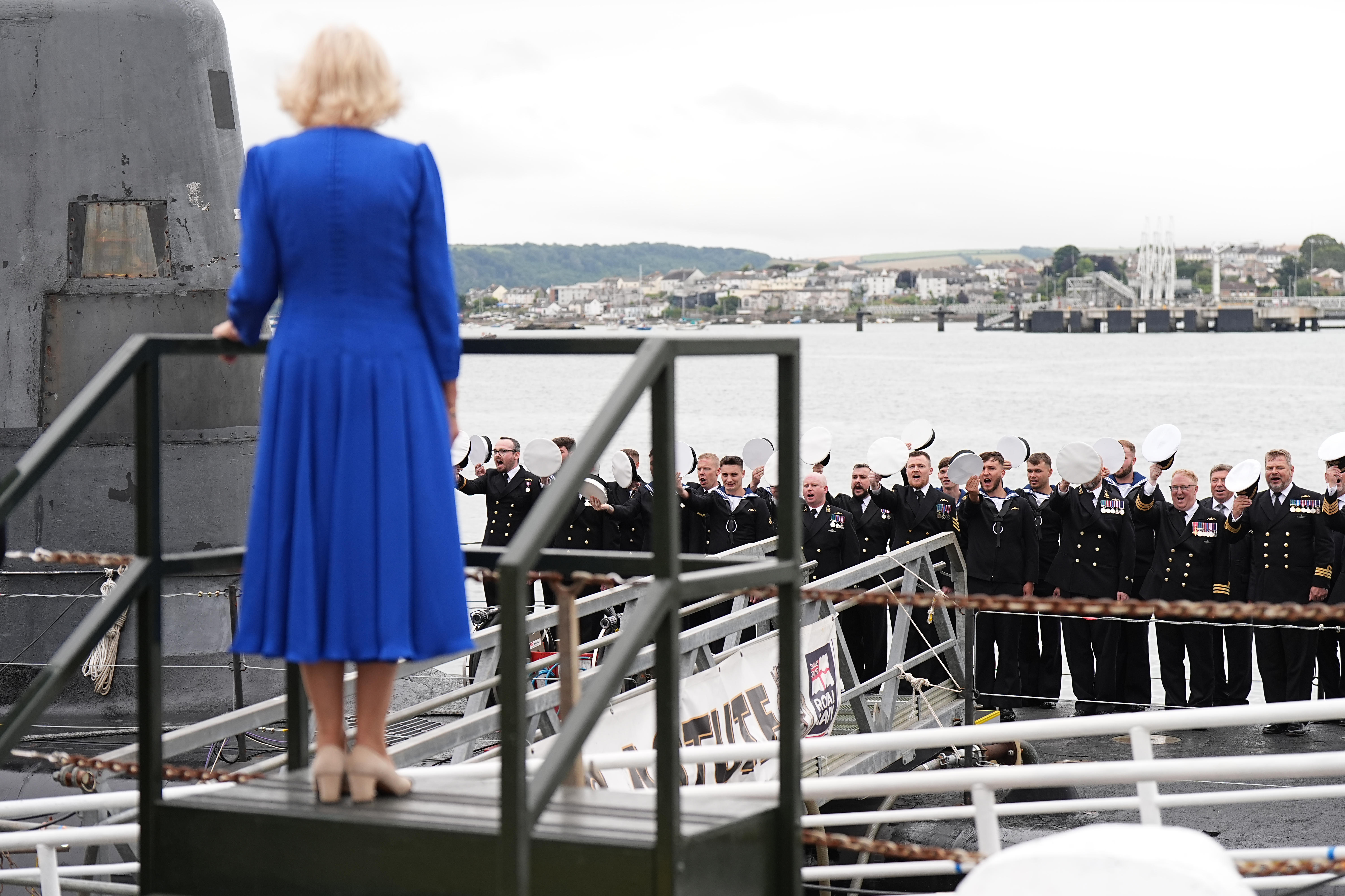 Queen Camilla wearing a blue dress standing with her back to the camera as naval officers take their hats off and salute her