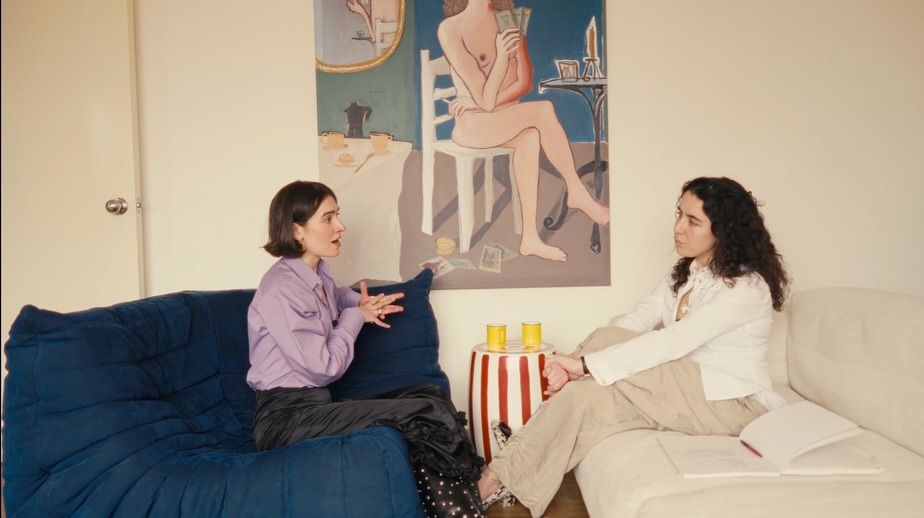 The interior of a young, brunette, curly-haired woman's living room-cum-kitchen, designed with pops of unexpected color, unique artwork, and sculptural sofas in blue and cream.