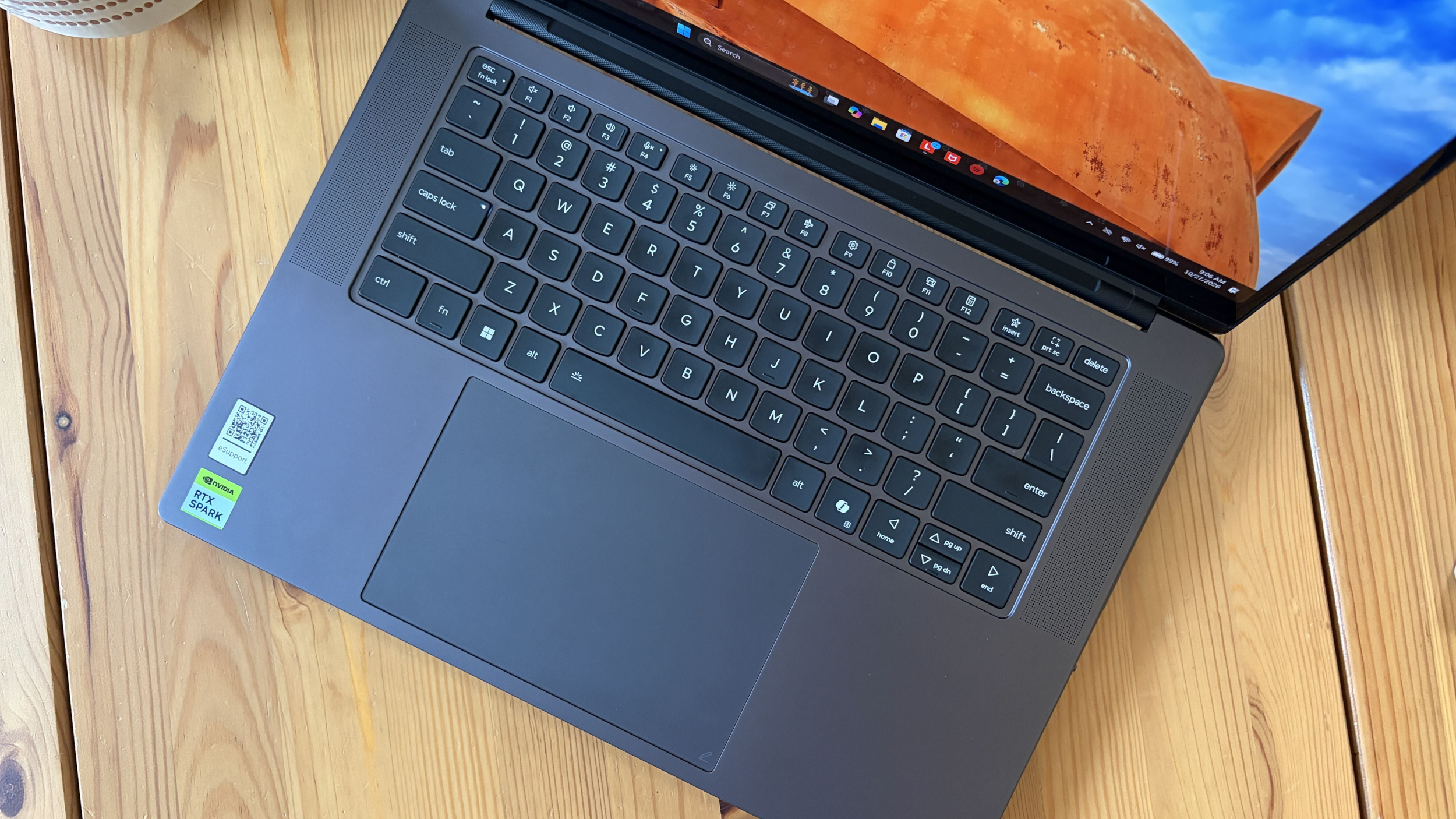 A dark gray laptop sits diagonally on a pale wooden table, its keyboard and touchpad visible beneath a screen showing orange earth and blue sky.