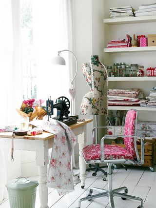 Sewing room with desk by window, tailor's dummy, open shelving with piles of fabrics and jars full of buttons and thread