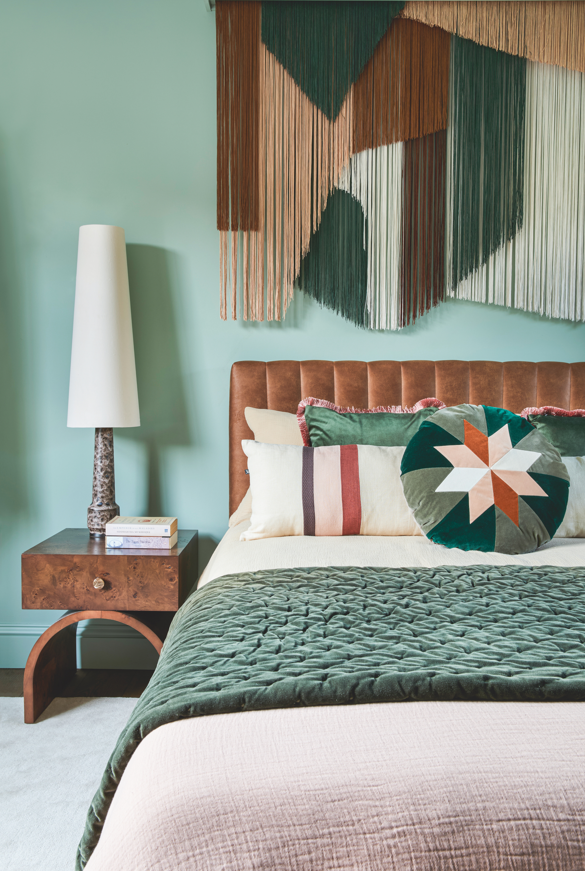 A turquoise colored bedroom with a tassel wall hanging above the bed, which has teal colored cushions and a dark wood nightstand to the left.