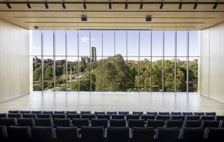Auditorium with black chairs closer to us, and panoramic windows that cover the entire far wall, through which we see the landscape outside.