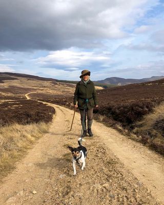 Queen Camilla walking down a dirt road holding a walking stick with her dog, Beth, running in front