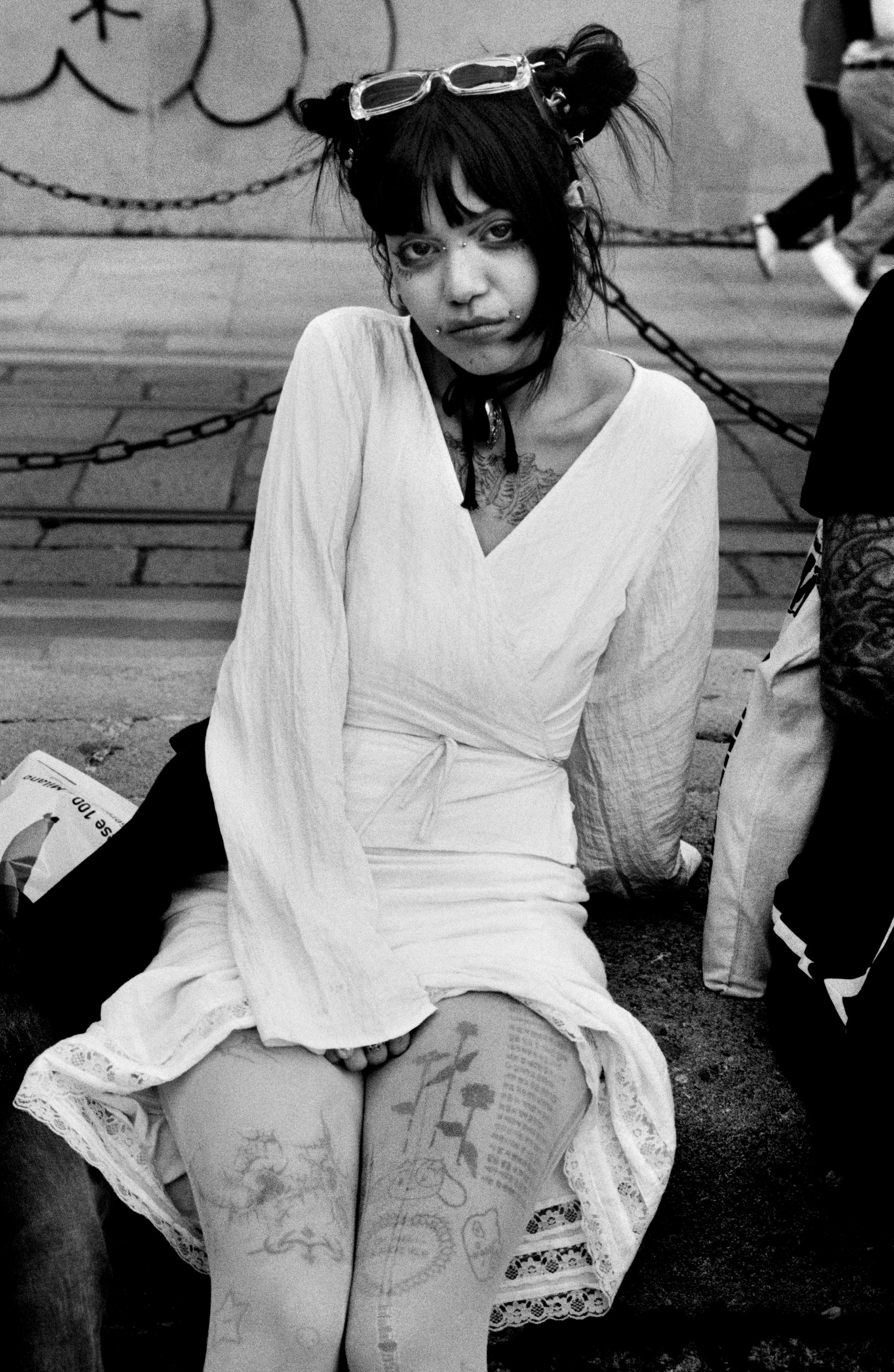 A young woman with dark hair in buns, facial piercings and tattooed legs sits on a stone ledge in a white wrap dress, gazing at the camera.