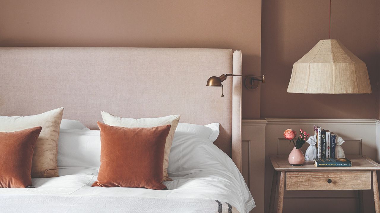 A light brown-painted bedroom with a pendant light hanging above the bedside table and rust-coloured velvet cushions placed on the bed