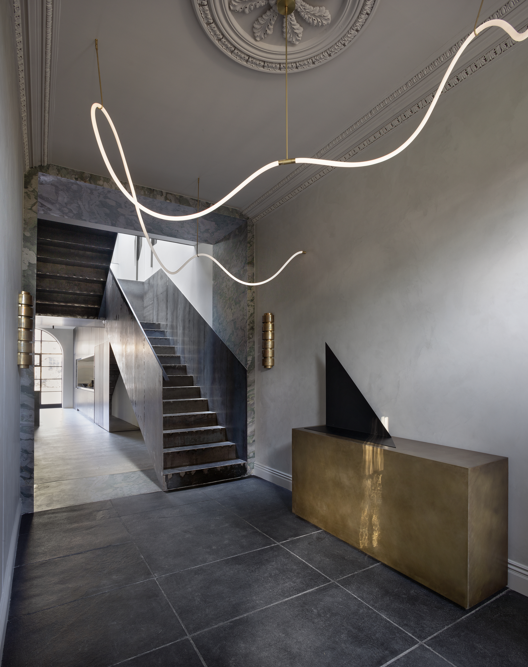 Image of a grand entryway with dark gray stone flooring, a gold, metal console, a stone doorway, and metal stairs.
