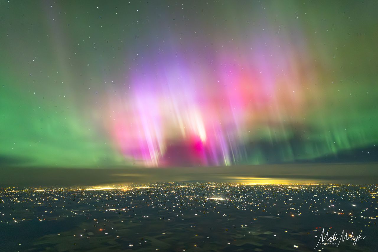 curtains of magenta and green light dance in the sky, yellow spots from city lights fill the scene below.