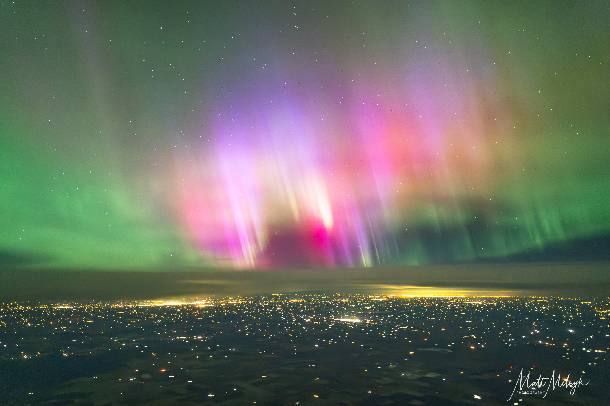 curtains of magenta and green light dance in the sky, yellow spots from city lights fill the scene below. 