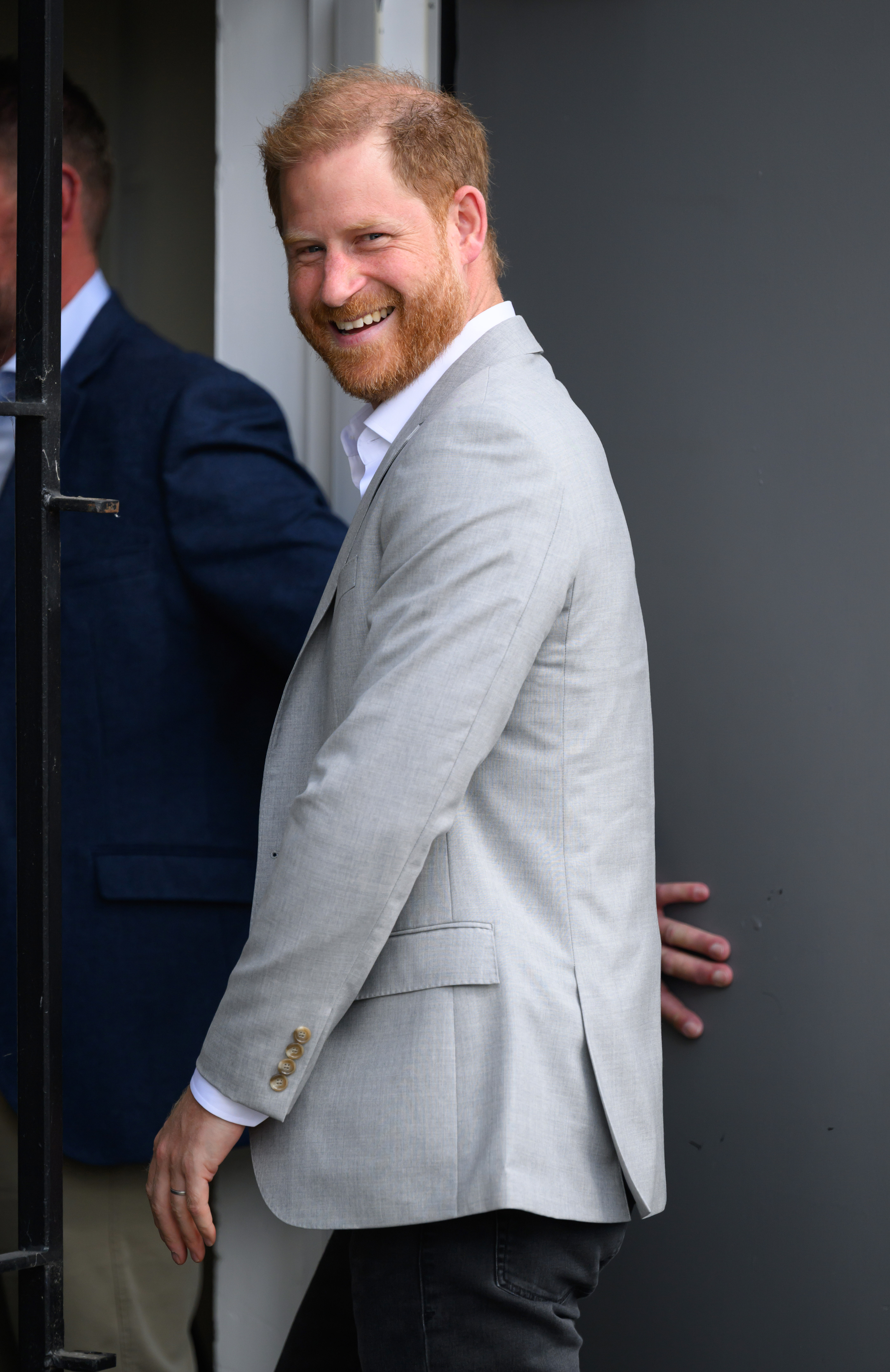 Prince Harry wears a gray suit jacket, a white shirt, and dark slacks and has red hair as he visits the UK in September 2025