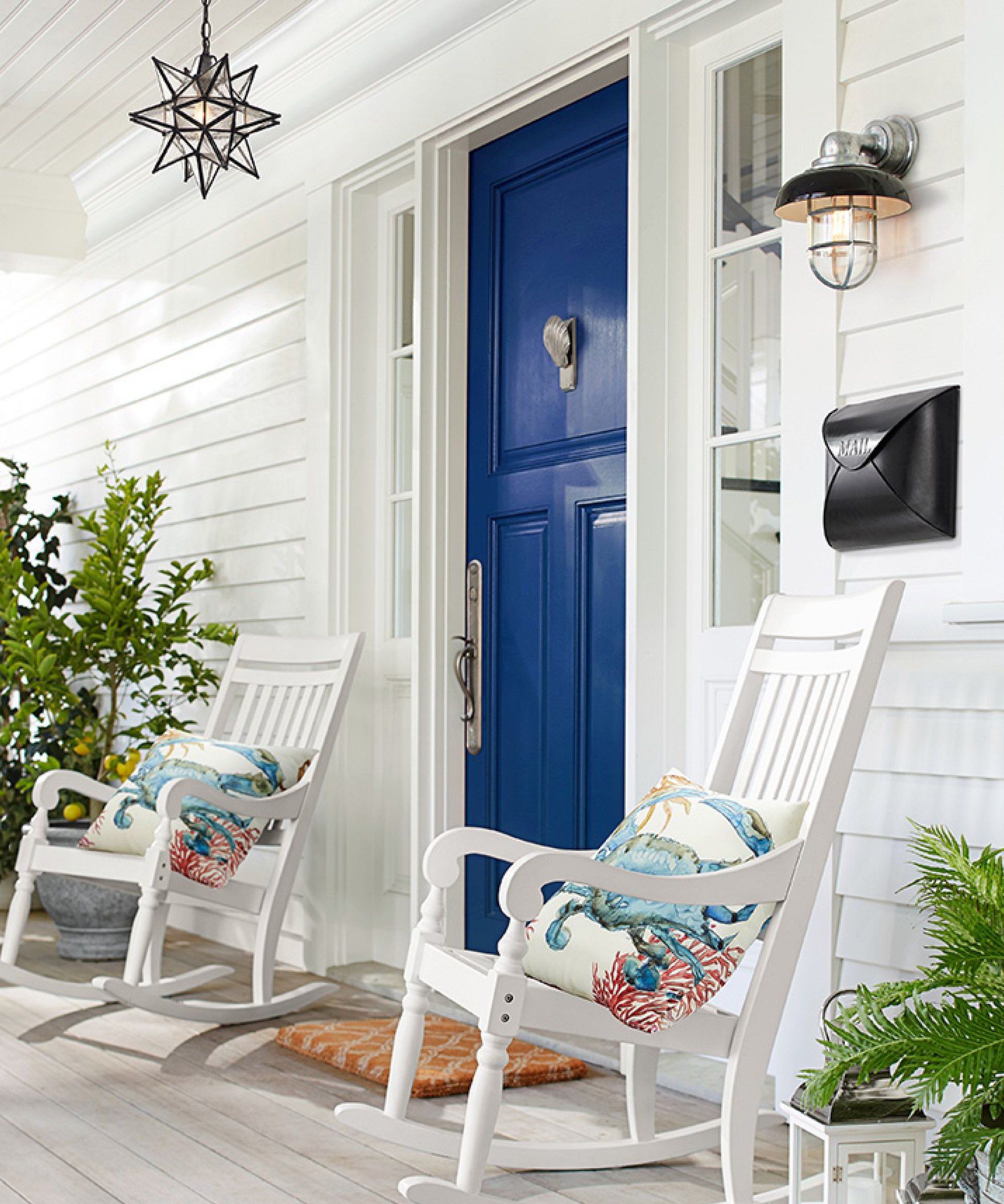 a blue front door with two white rocking chairs on either side with decorative pillows on them, a black mailbox, a wall sconce, and a star-shaped chandelier