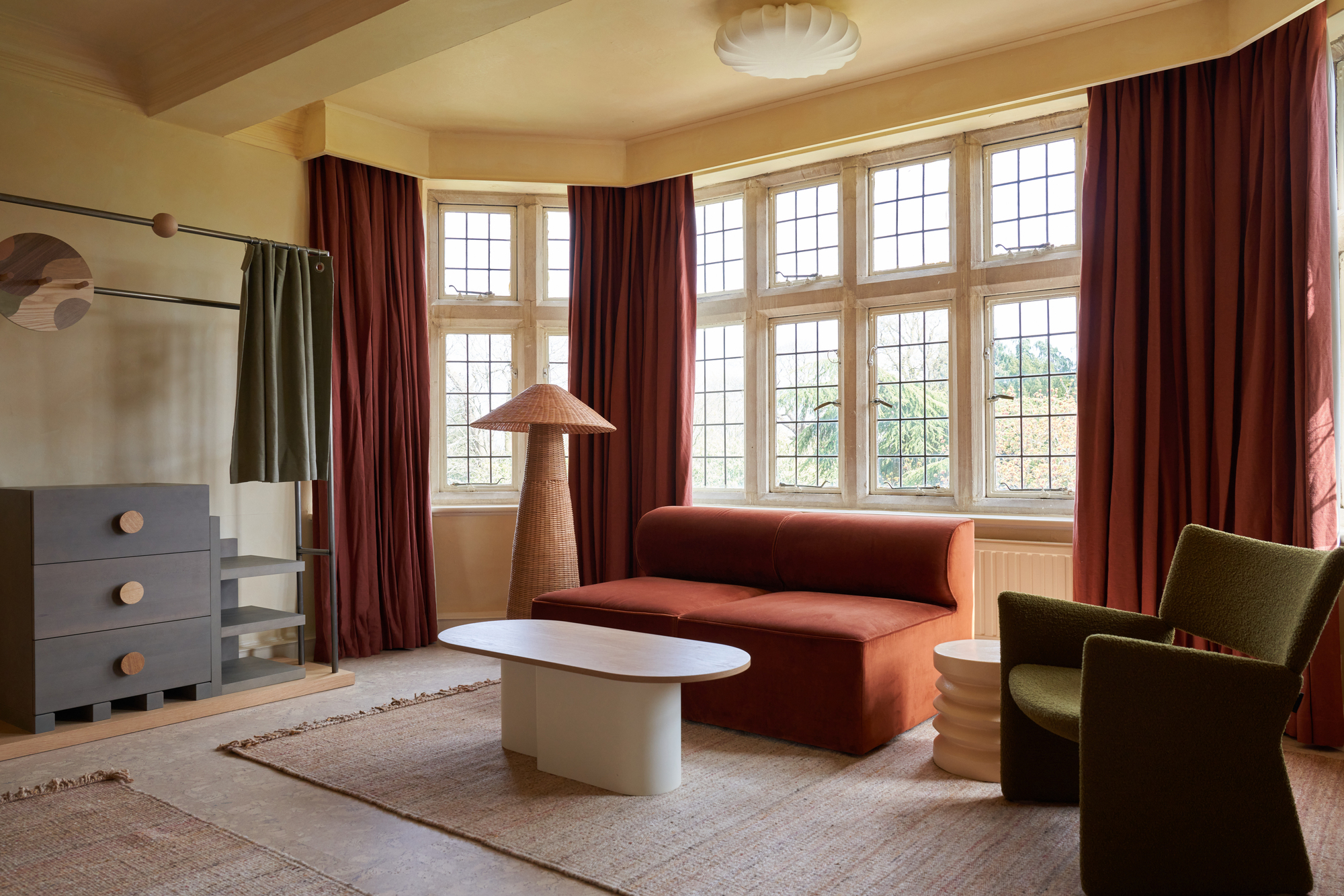 hotel bedroom with orange sofa, green armchair, white coffee table, jute rugs, large windows with red drapes, a ferm living rattan floor lamp, freestanding wardrobe, and yellow ceiling