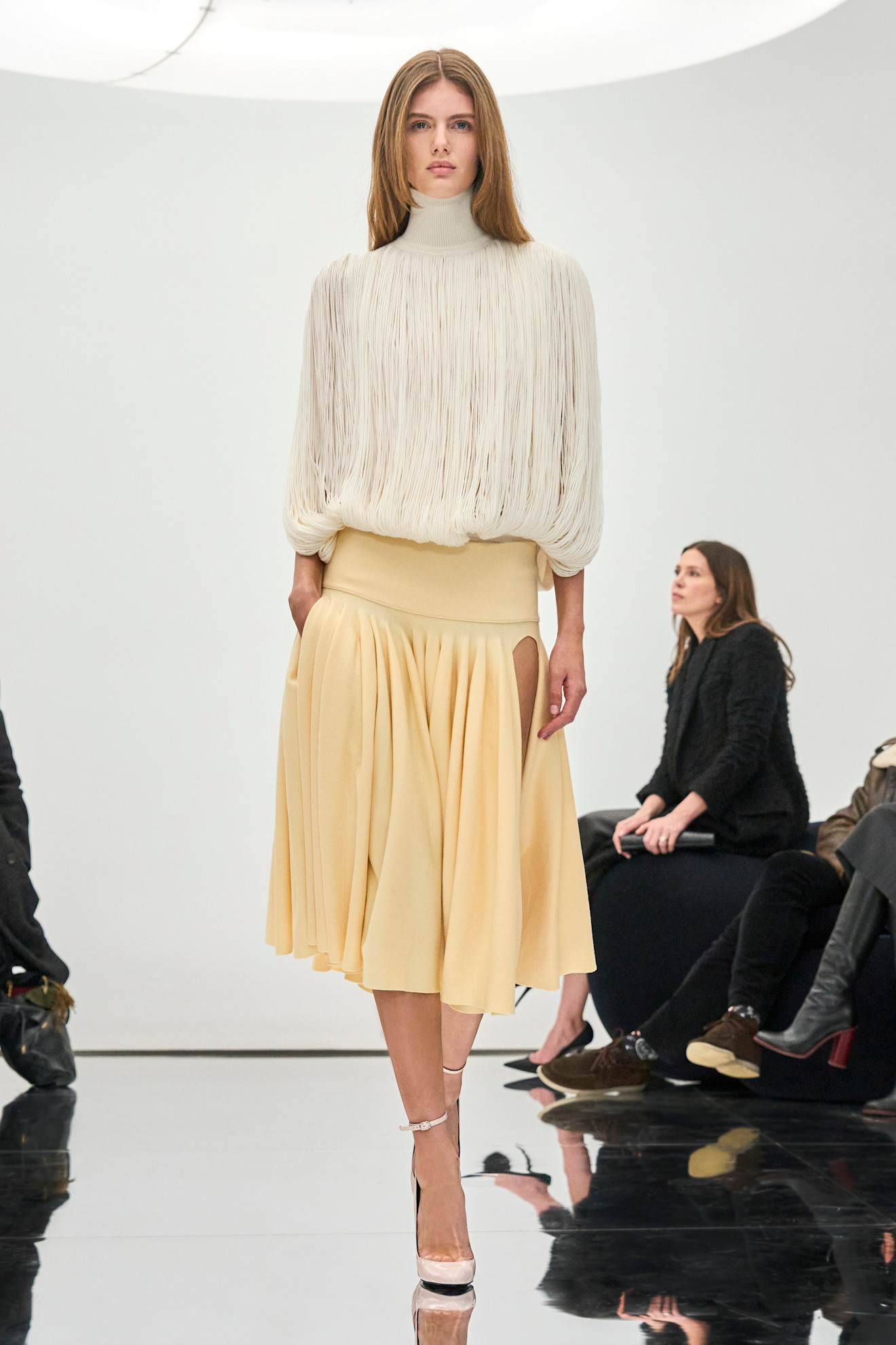 Photo from the F/W 24 Alaïa show of a dirty blonde model wearing a high-neck butter yellow fringe top with a drop-waist yellow skirt and blush pink round-toe heels.