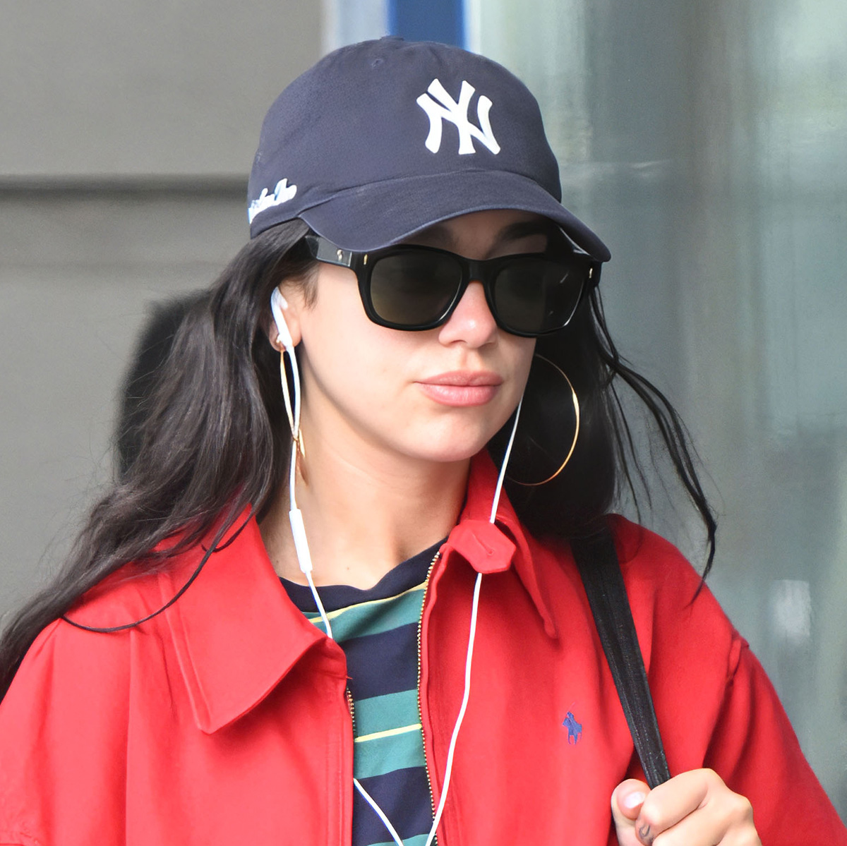 Dua Lipa at JFK wearing a red Polo Ralph Lauren cotton jacket with a retro striped tee, washed-black jeans, black leather boots, and a NY Yankees baseball cap