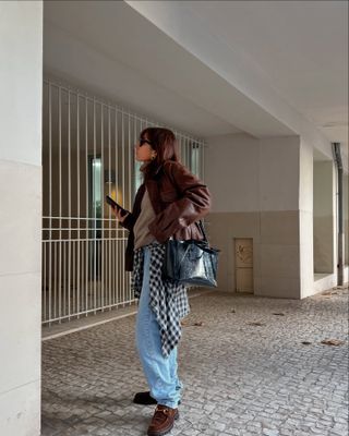 Fashion influencer @emilie_tla wearing a burgundy leather jacket, faded blue jeans, and trendy suede loafers..