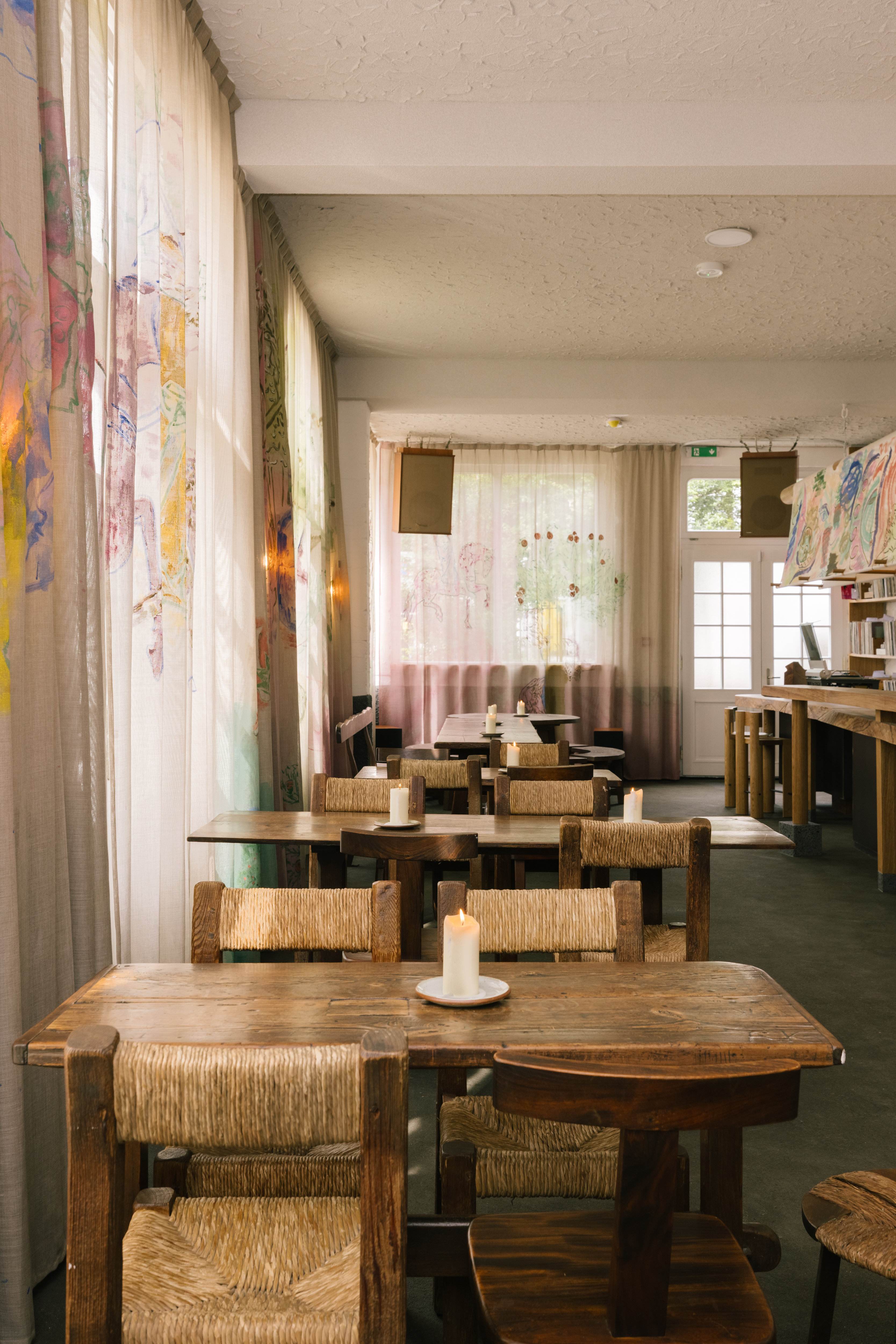 A naturally lit restaurant dining room with lived wood and rattan table sets, mural curtains and a mural frieze, and bare cement floors and ceilings, the latter of which painted white.