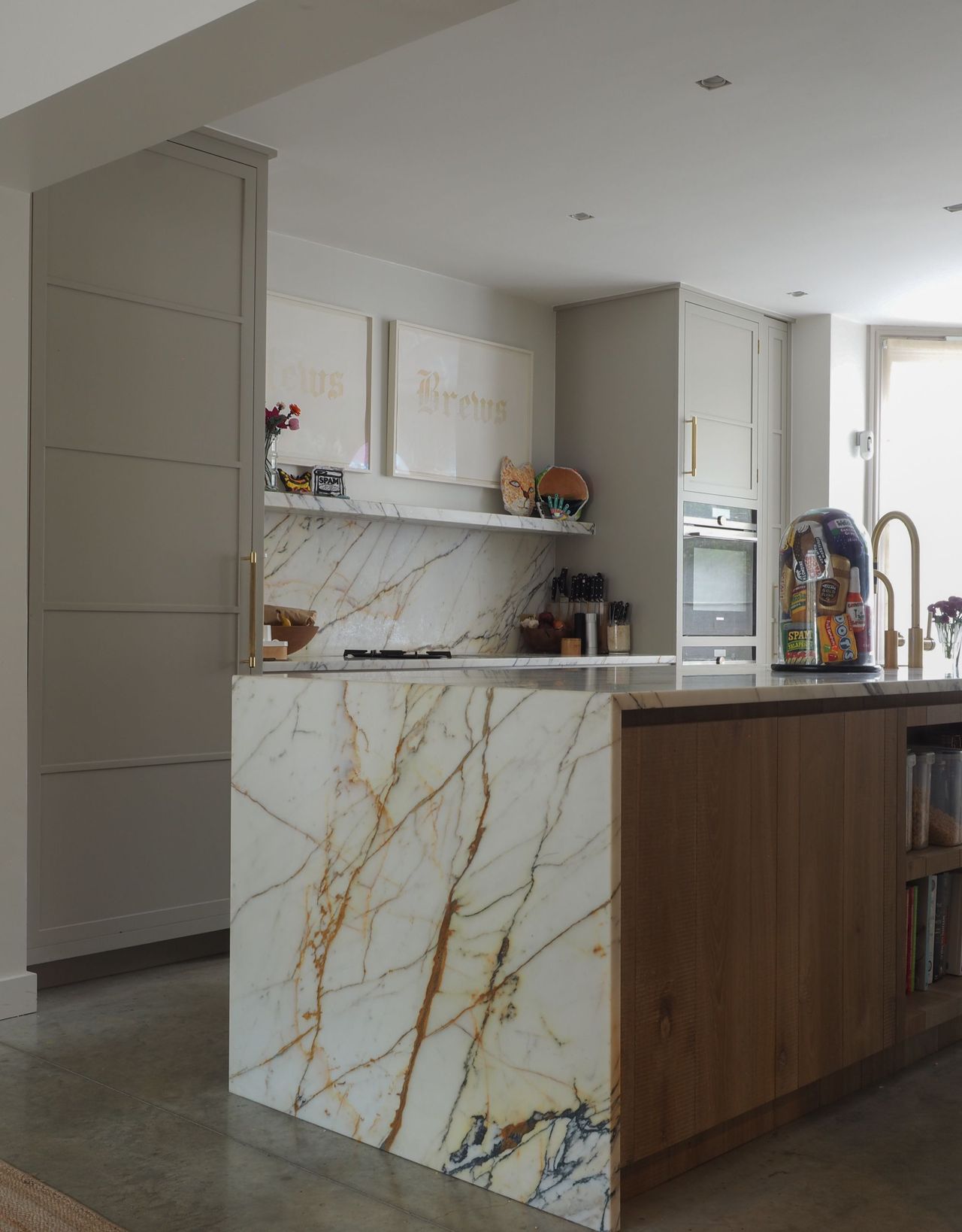 A modern kitchen with a large kitchen island with wooden cabinets and open shelves with a marble top, and artwork on the wall and in a cloche on top of the island