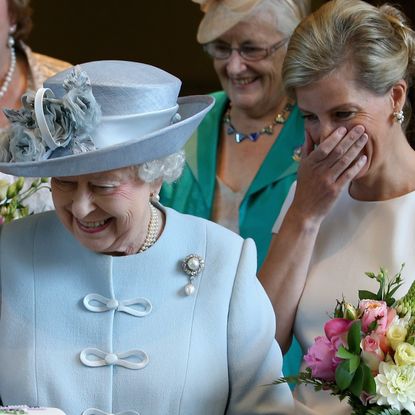 Queen Elizabeth wears a light blue suit and matching hat and everyone laughs as she cuts a cake