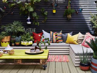 A backyard area with decked floor surface, horizontal wood fence paneling decor with black paint decor, grey seating area with assortment of colorful outdoor cushions and yellow painted outdoor table with castor wheels