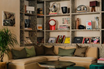 A collectibles-filled hotel lounge with a beige, velvety sofa covered in brown and green pillows, acrylic and wooden coffee tables, a patterned floor topped by a velvety carpet, a paper lantern, and plants.