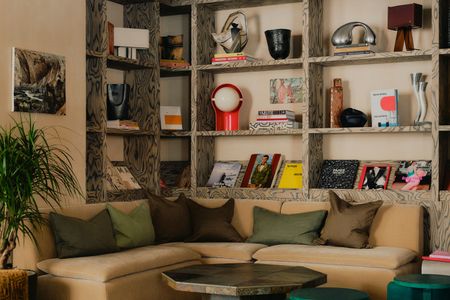 A collectibles-filled hotel lounge with a beige, velvety sofa covered in brown and green pillows, acrylic and wooden coffee tables, a patterned floor topped by a velvety carpet, a paper lantern, and plants.