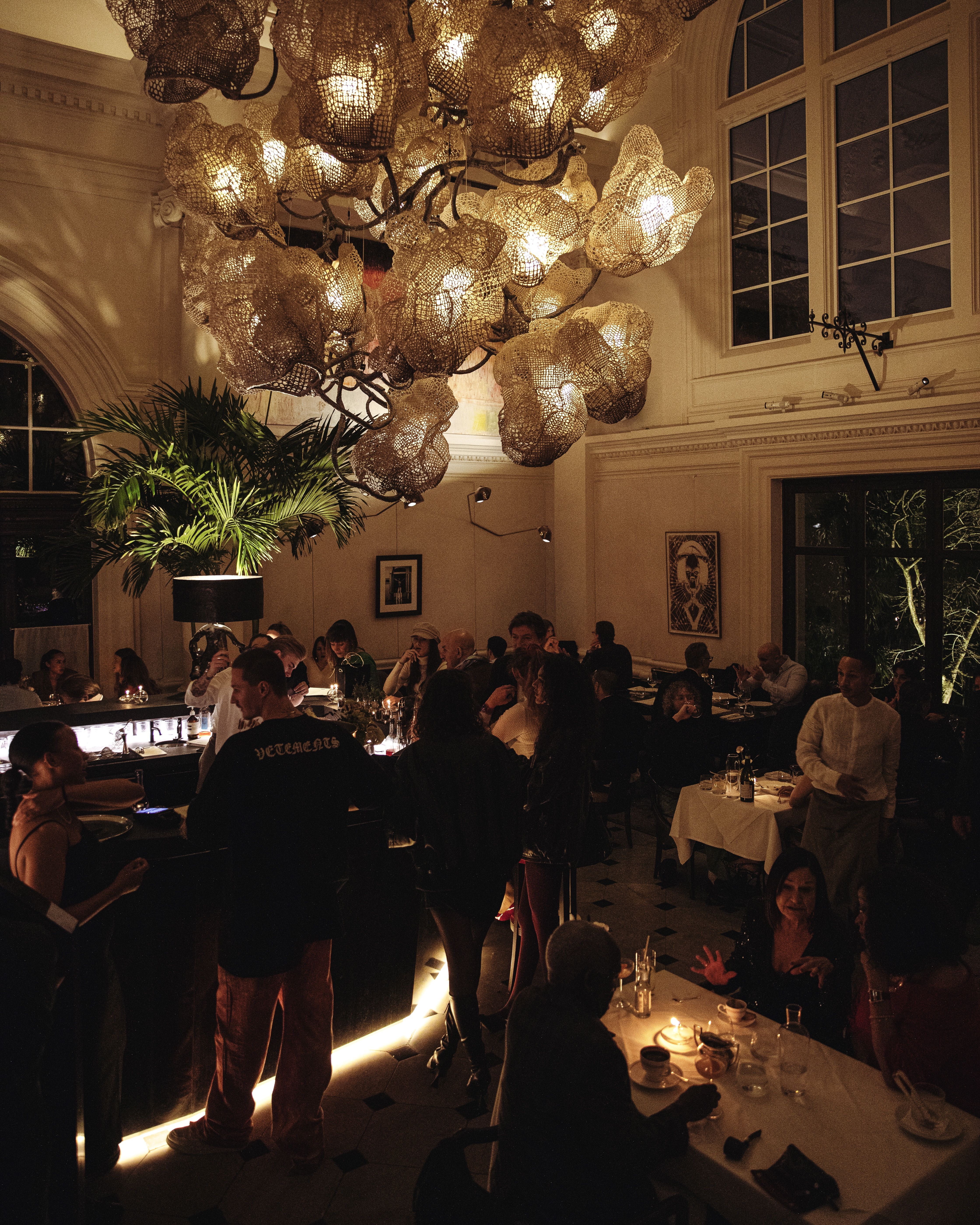 A nightlife venue with a golden, dimly lit atmosphere features a sculptural chandelier made of tree branches-looking metallic parts, lots of people dancing or eating at bistro-style tables, a DJ playing, and plants.