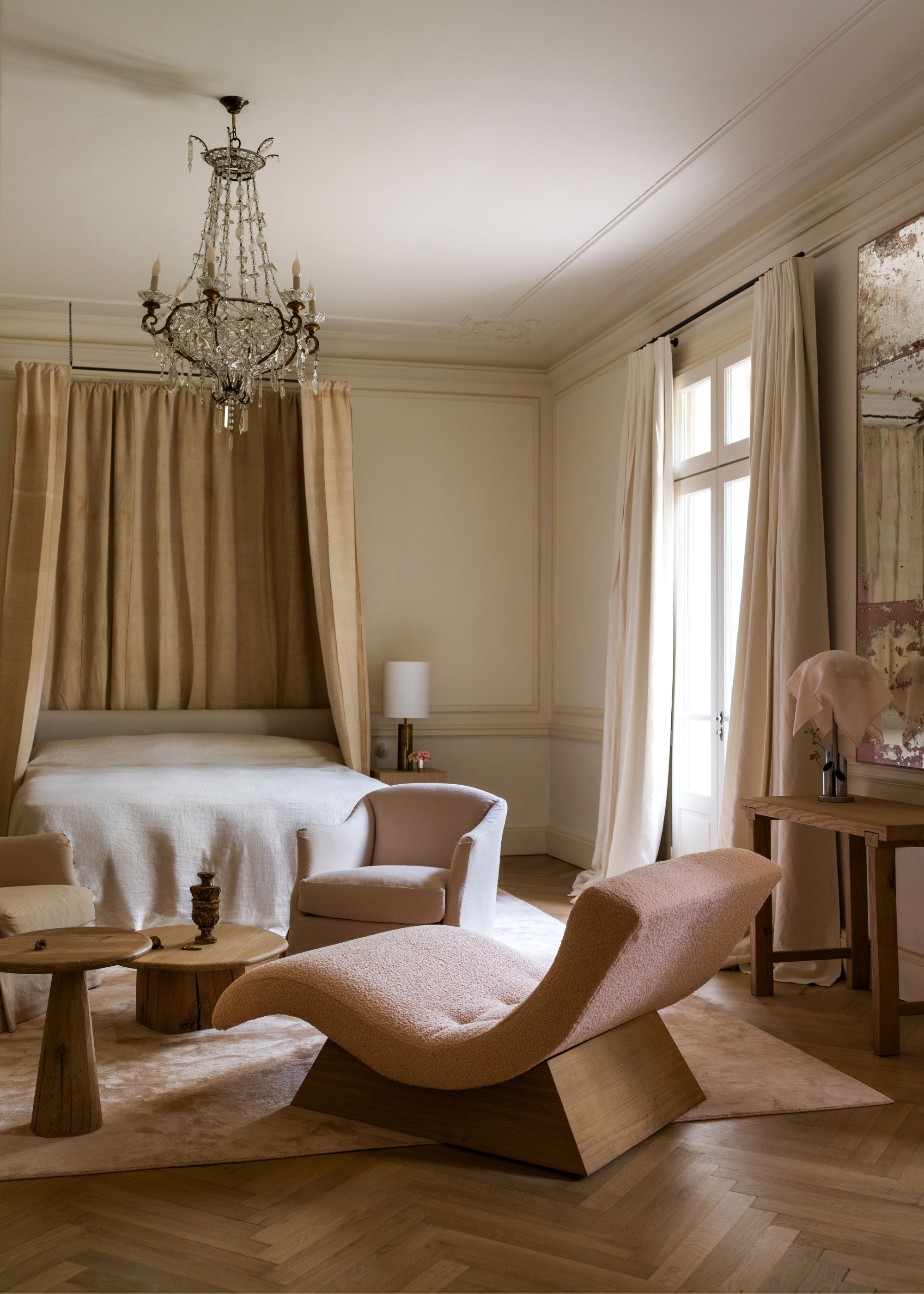 A bedroom with quiet luxury design, including a chaise lounger, wood side tables, a bed with a draped canopy, a chandelier, and vintage mirrors