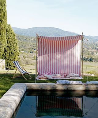 Backyard ideas on a budget, with a red and white fabric draped over posts to create a shaded spot beside a stone pool, with a hilly backdrop.