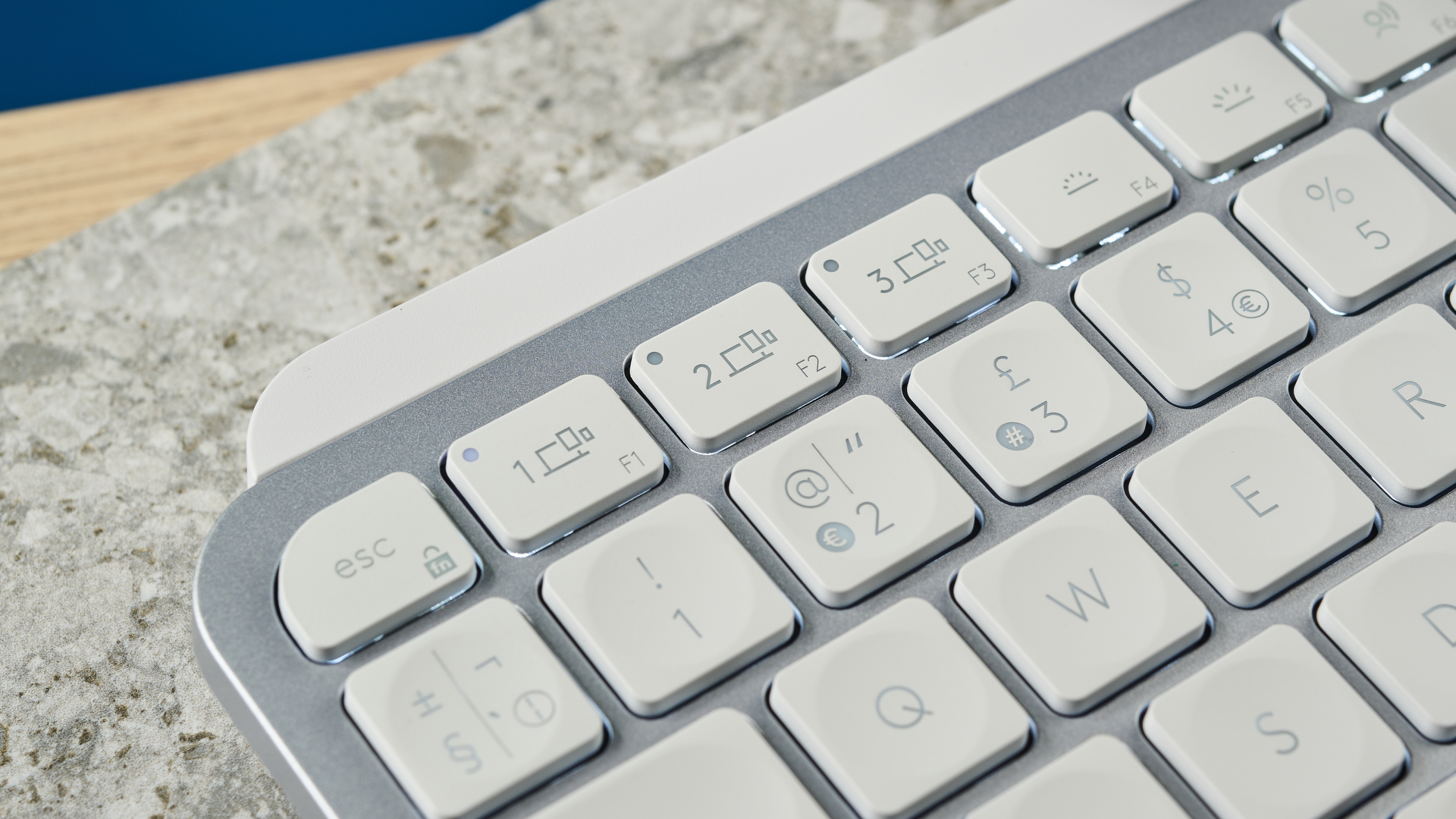 A photograph of the Logitech MX Keys Mini in light gray, positioned on a stone slab with a blue wall in the background.