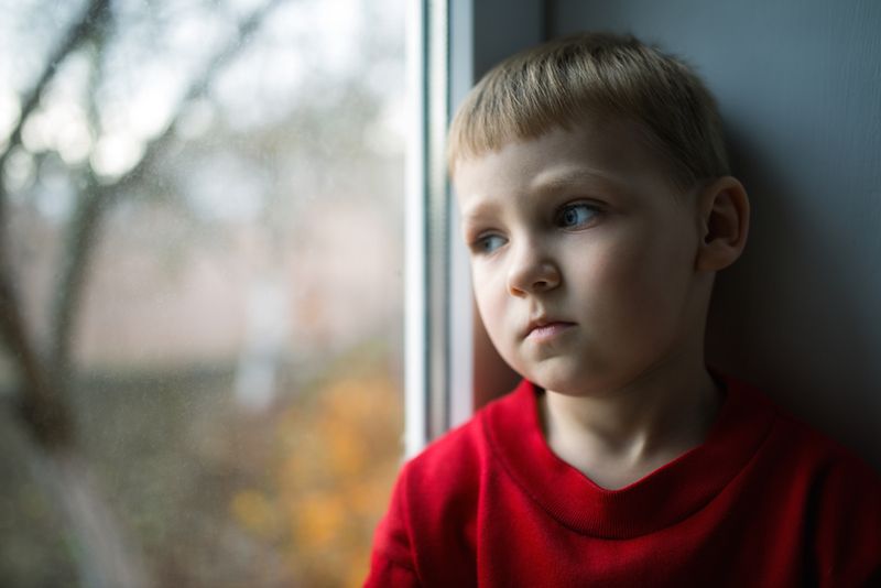 A young boy looks out a window, looking very sad.