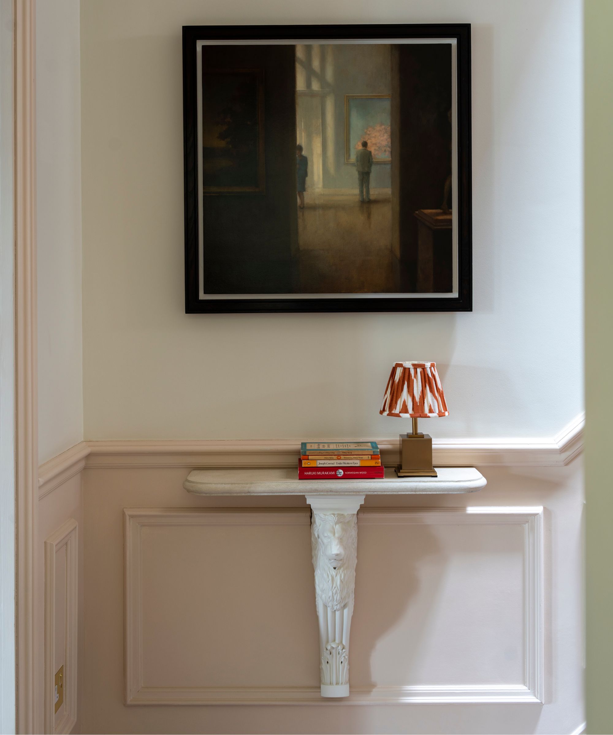 A white and pale pink stairway with white shelving with a stack of books and a small table lamp with a red and white lampshade. Above is a framed artwork in a black frame.