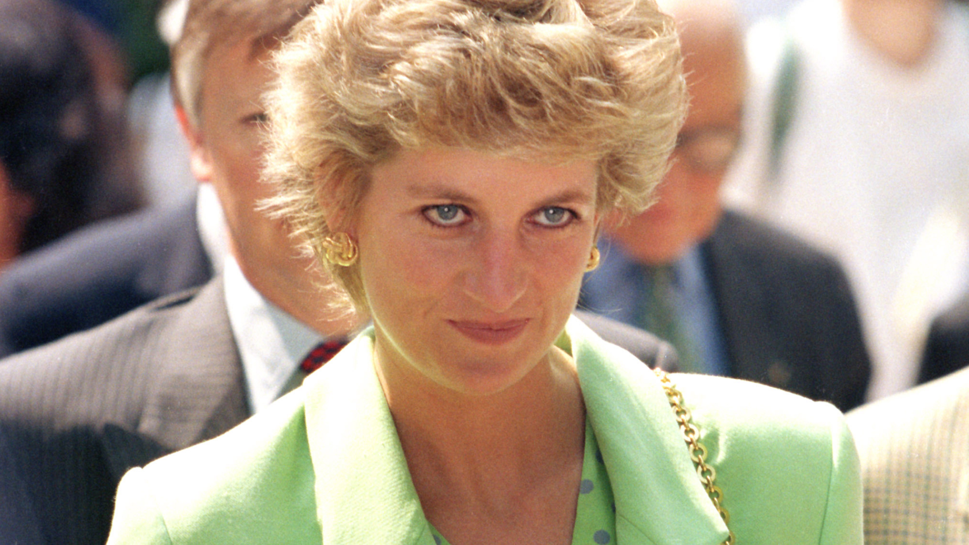 Princess Diana wears a bright green jacket with a matching polka-dot shirt underneath and has short blonde coiffed hair, a bag with a gold chain strap, and she's wearing black eyeliner