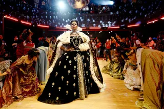 A woman (Dominique Jackson as Elektra), wearing a Victorian queen's gown, stands in a ballroom as a crowd dressed in a period wardrobe bows to her, in 'Pose.'