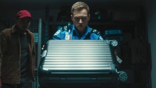 Taron Egerton lifting a carry-on suitcase up with a man in a red baseball hat behind him.