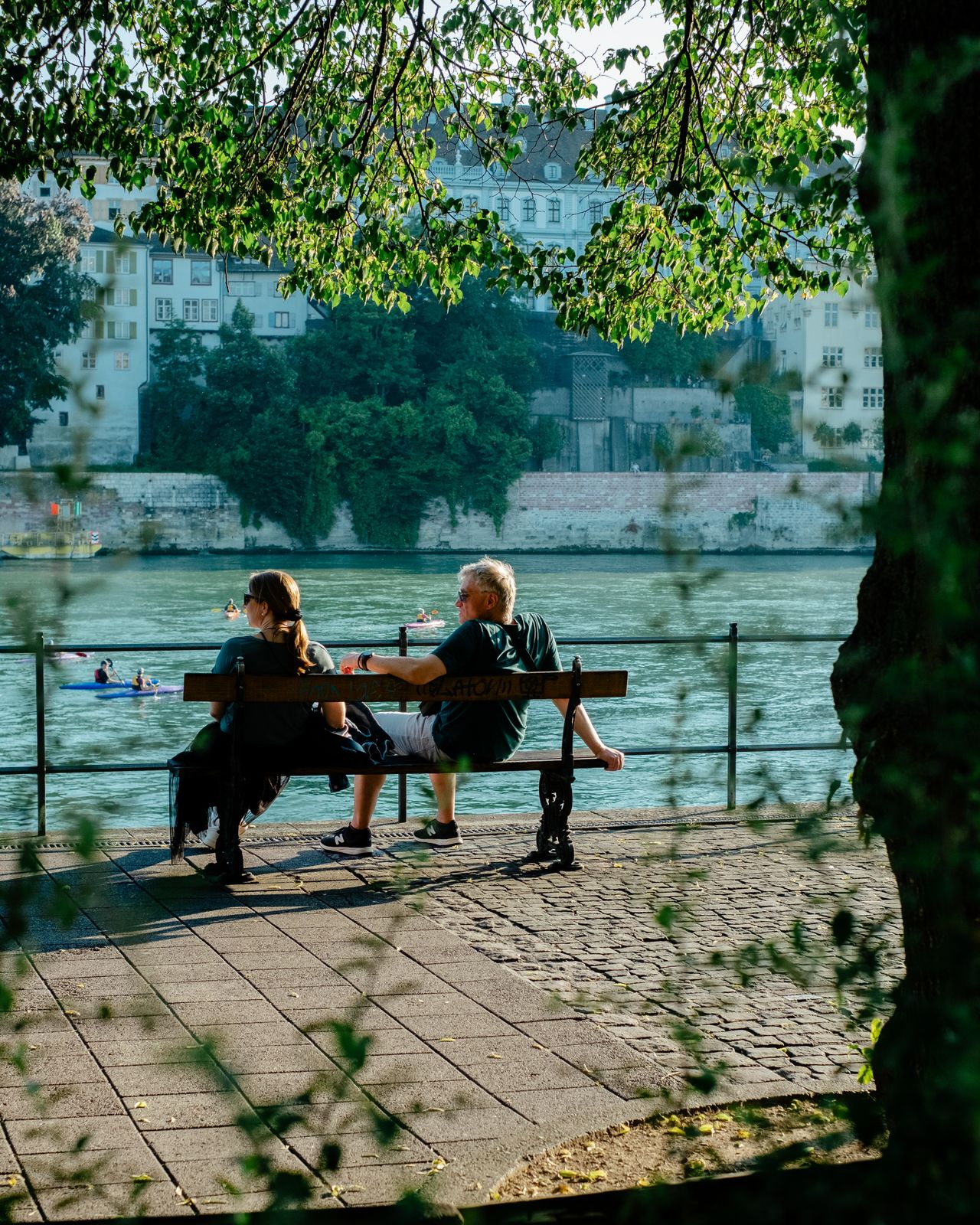 A ypung woman dressed in a black bottoned-up top and a beige, pleated skirt smiles at the camera while standing in front of a river canal bathing in sunshine, while in another image, two middle-aged people sit on a bench on the same, leafy canal.