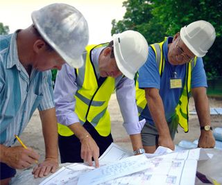 three men stood in row wearing hi vis jackets, white hard hats and looking at plans on building site