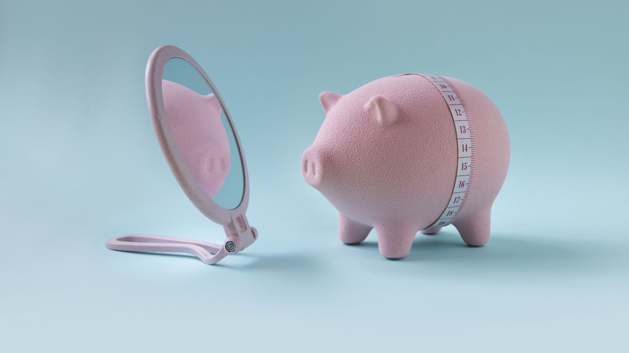 A piggy bank with a measuring tape around its belly appears to be looking in a hand mirror.