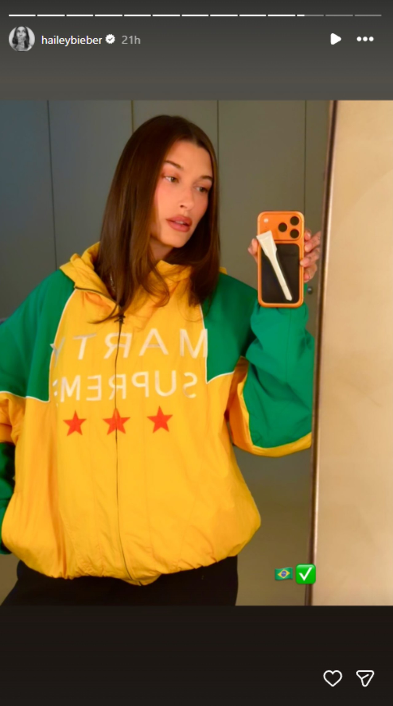 Hailey Bieber taking a mirror selfie while wearing a yellow and green Marty Supreme jacket that has the colors of Brazil's flag.