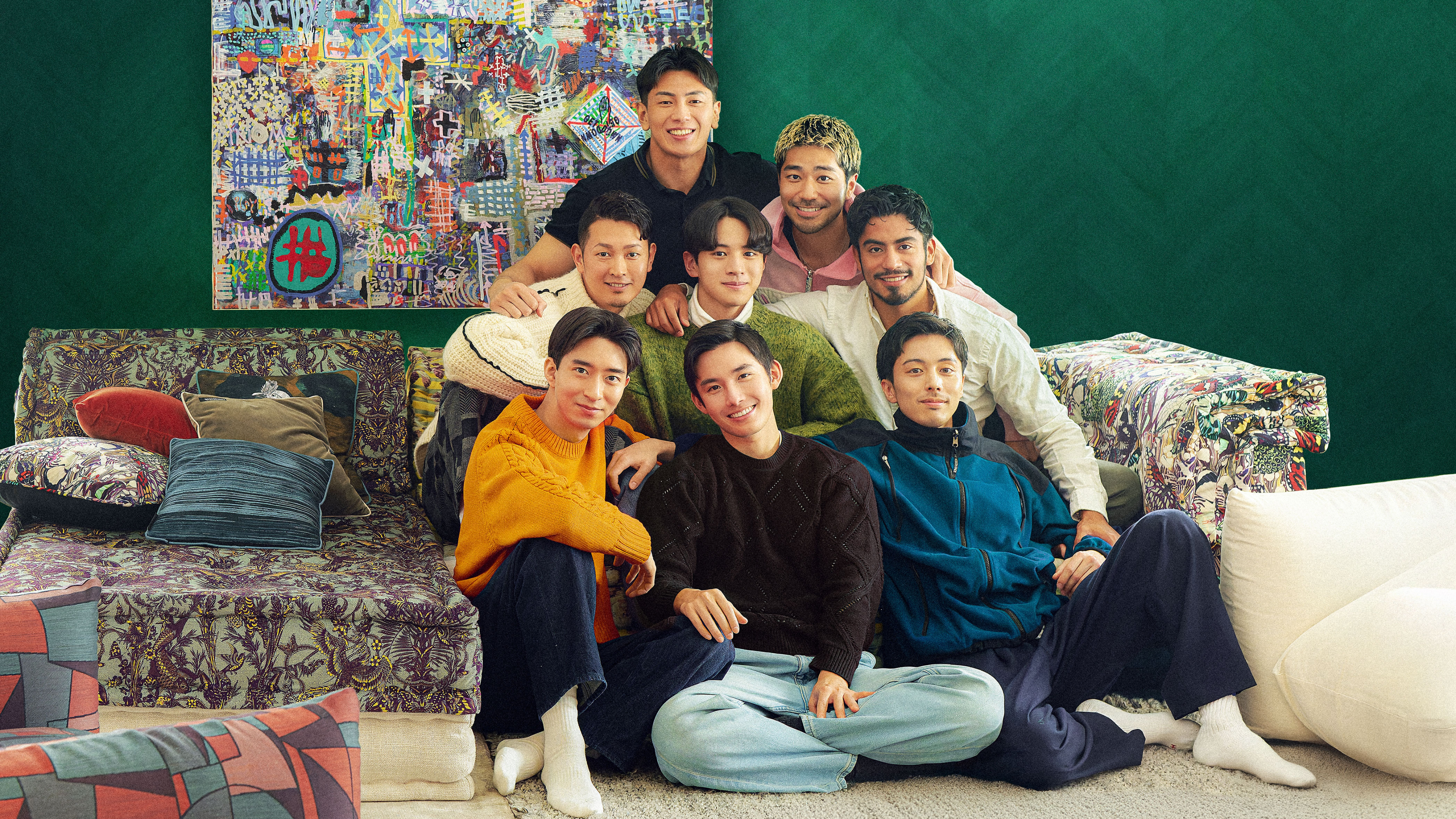 Eight Asian men sit in a pyramid formation in front of a printed sofa, multicolored painting, and green wall, in the group portrait for the cast of Netflix's 'The Boyfriend' season 2.