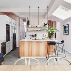 A modern kitchen-lounge with pink wall paint, trio of white pendant light fixtures and wooden island, built-in white cabinetry and oven appliance