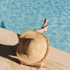 A pale girl with a large hat is sitting on the edge of a pool, possible wearing the best fake tan for pale skin